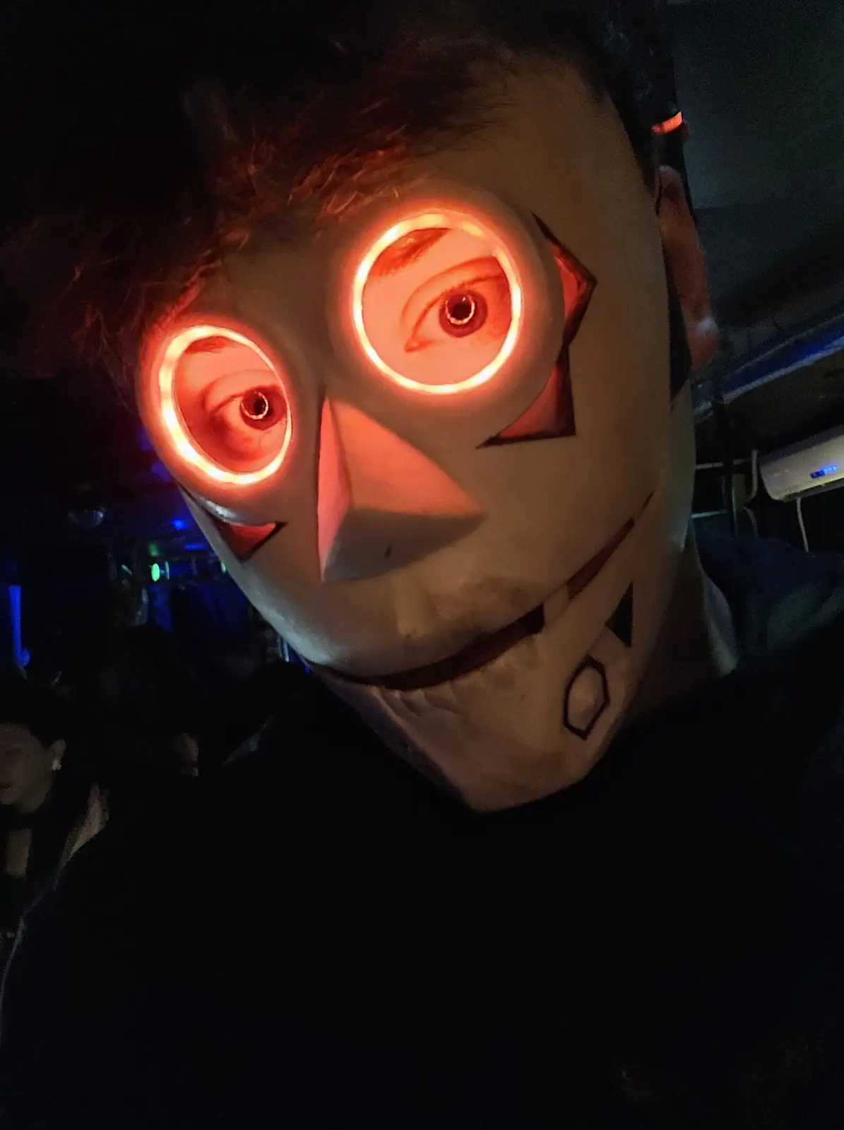 A person wearing a white, cartoonish mask with glowing red circular eye holes, revealing their actual eyes inside. The mask…
