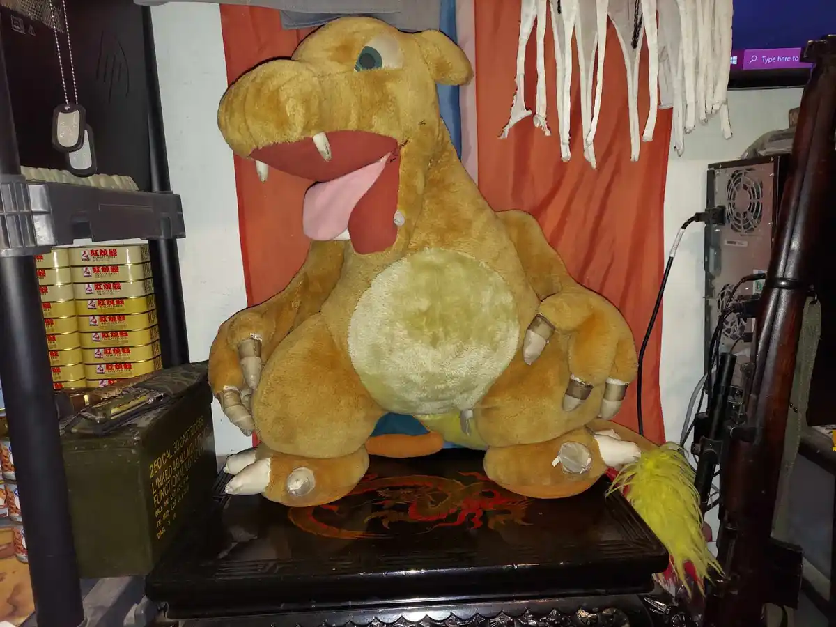 A large, tan plush dragon toy with a wide-open mouth, pink tongue, and white claws sits on a black ornate table. Its claws…