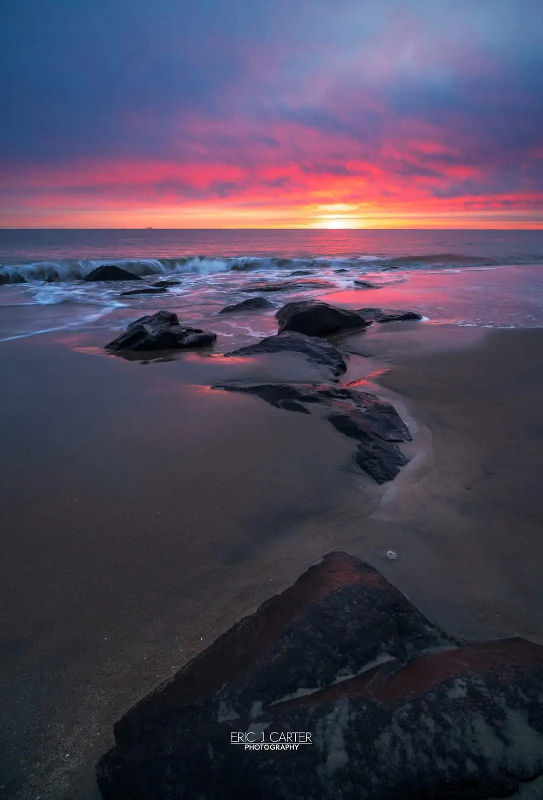 A serene beach scene at sunset with vibrant pink, orange, and purple clouds reflecting on the water and wet sand, featuring…