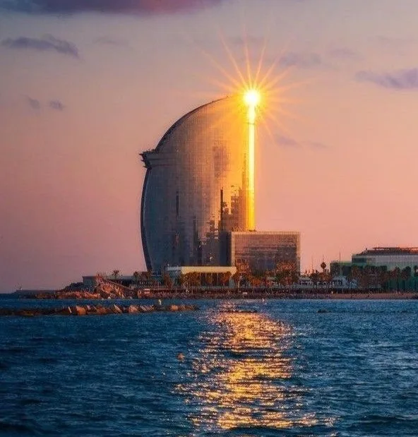 a scenic photo of the W Hotel in Barcelona at sunset, where the sun appears to shine directly through a gap in the…