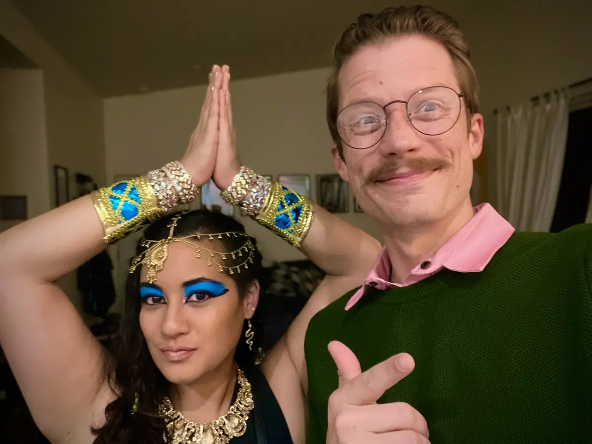A man in a green sweater and pink collared shirt with glasses and a mustache gives a peace sign while smiling at the camera…