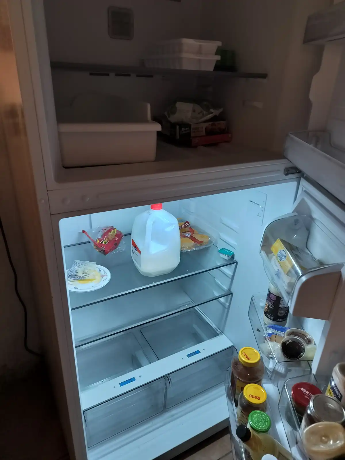 An open refrigerator with mostly empty shelves, containing only a gallon of milk, a plate with leftover food, and a few…