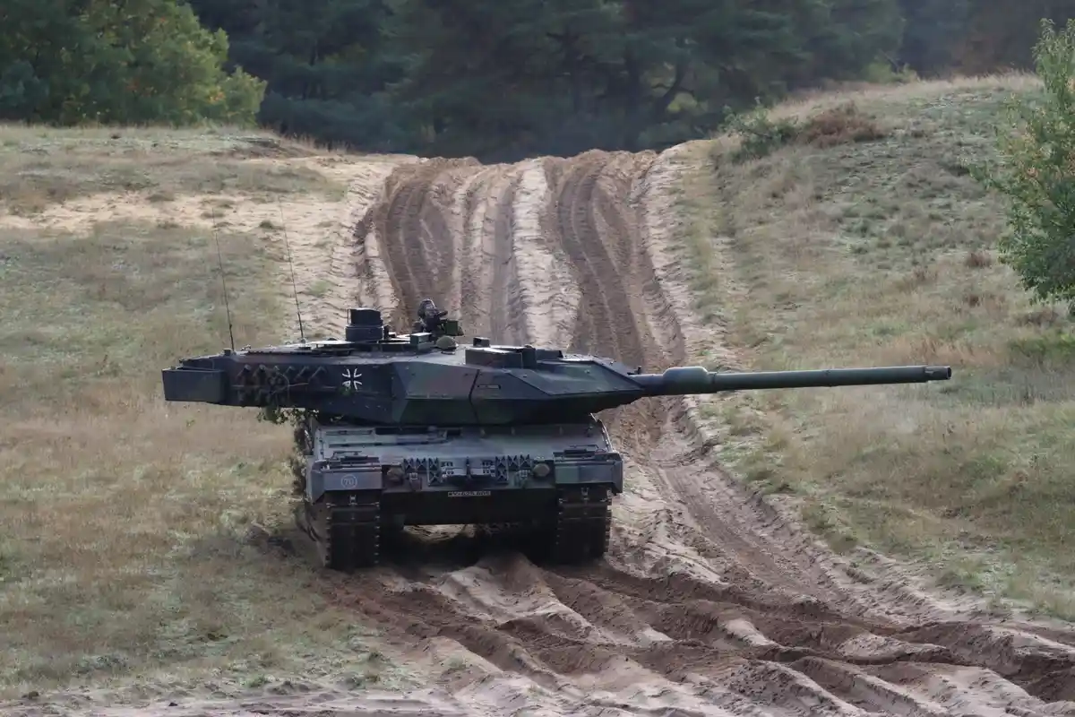 A German Leopard 2 tank, marked with the number 70 and a white cross, is shown driving down a muddy, rutted dirt track in a…