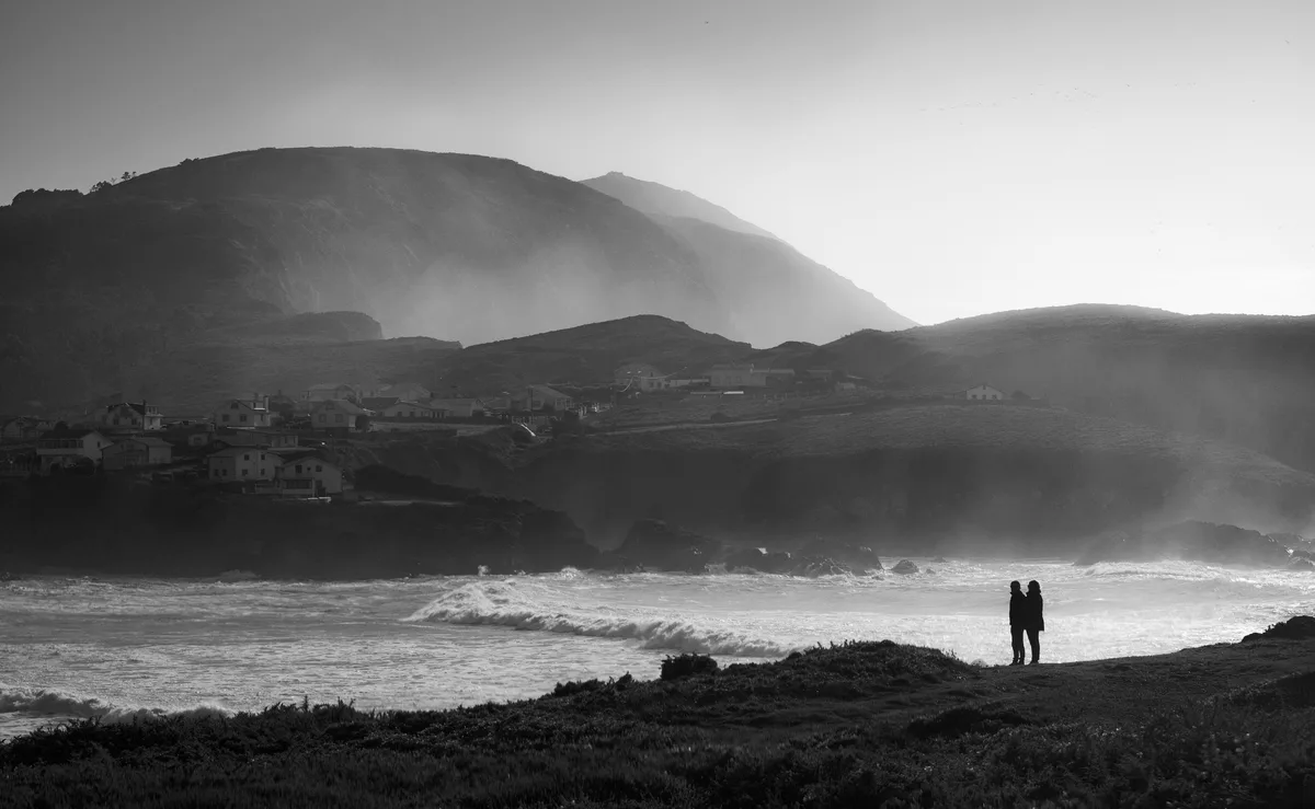 A black-and-white photo shows two silhouetted people standing on a grassy cliff overlooking a crashing ocean with waves and…