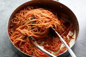 A close-up photo shows a large metal pan filled with cooked spaghetti in red tomato sauce, with two metal tongs resting in…