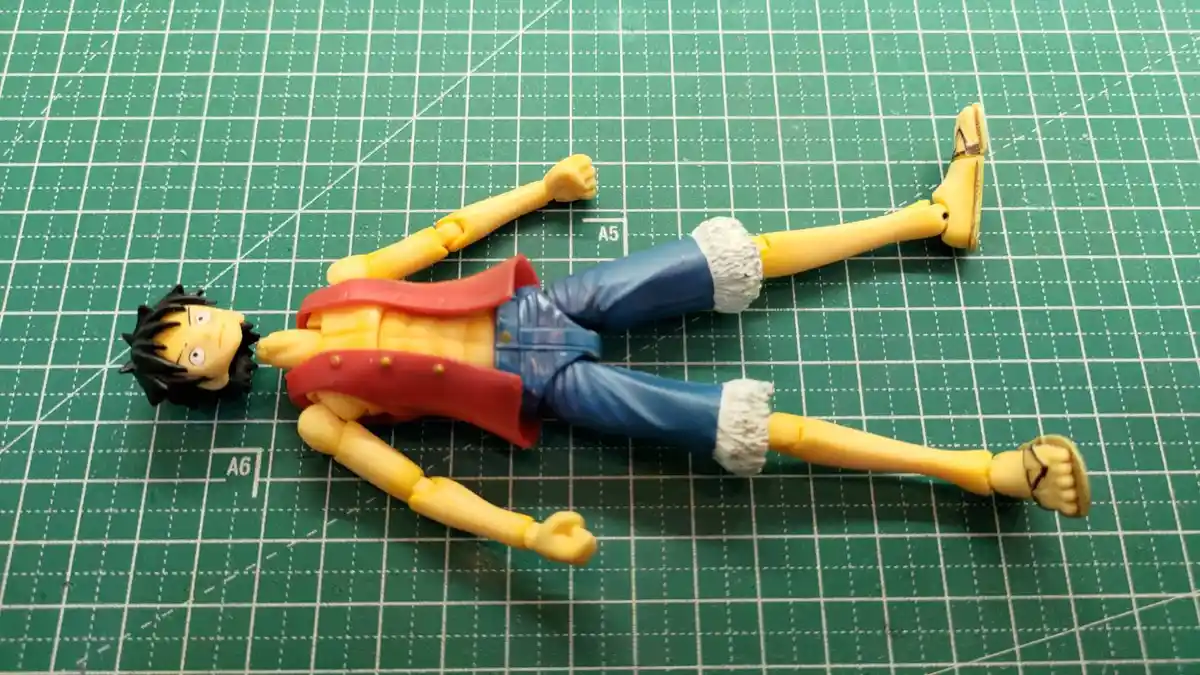 A plastic action figure of the anime character Monkey D. Luffy from One Piece lies flat on its back on a green cutting mat…