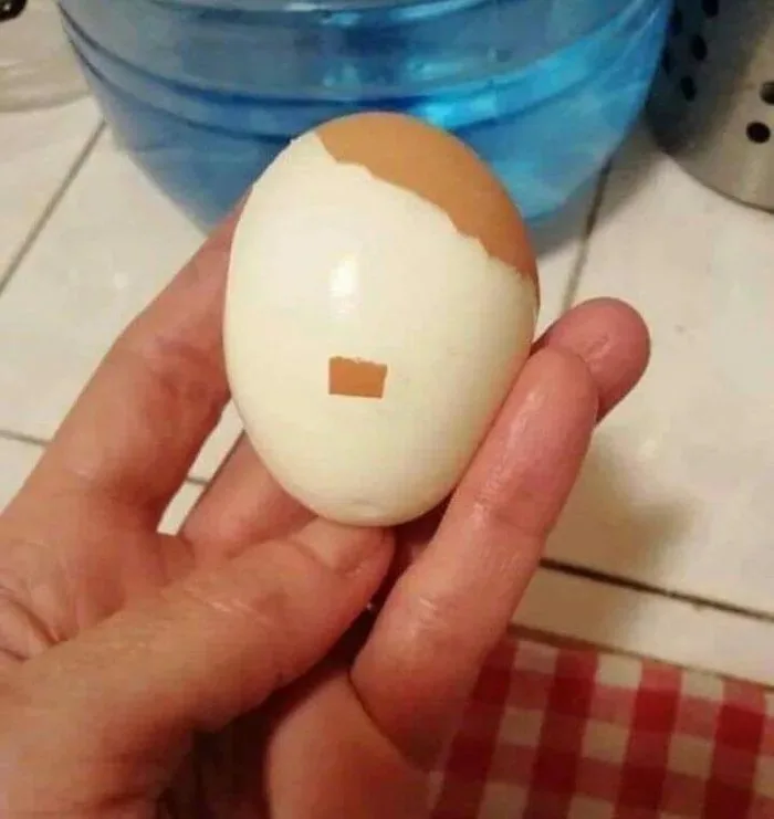 A hand holds a peeled hard-boiled egg with a small square of brown shell still attached to its white surface, resembling a…