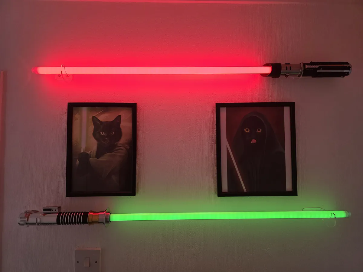 Two horizontal lightsabers, one red and one green, are mounted on a wall above two framed pictures of black cats. The left…