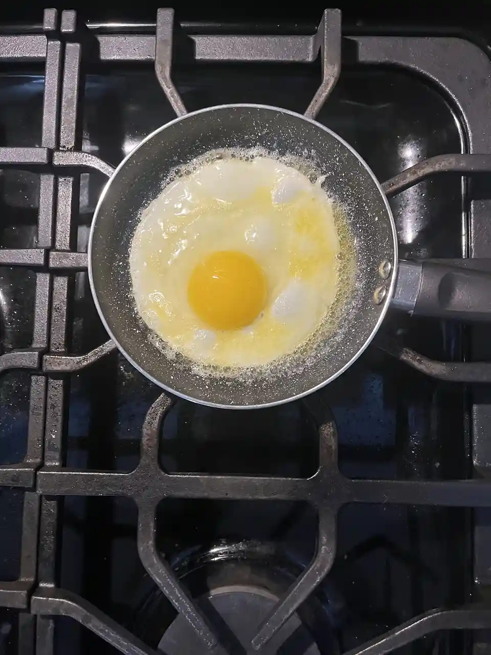 An overhead shot shows a single sunny-side-up egg frying in a small metal pan on a black gas stove grate. The egg white is…