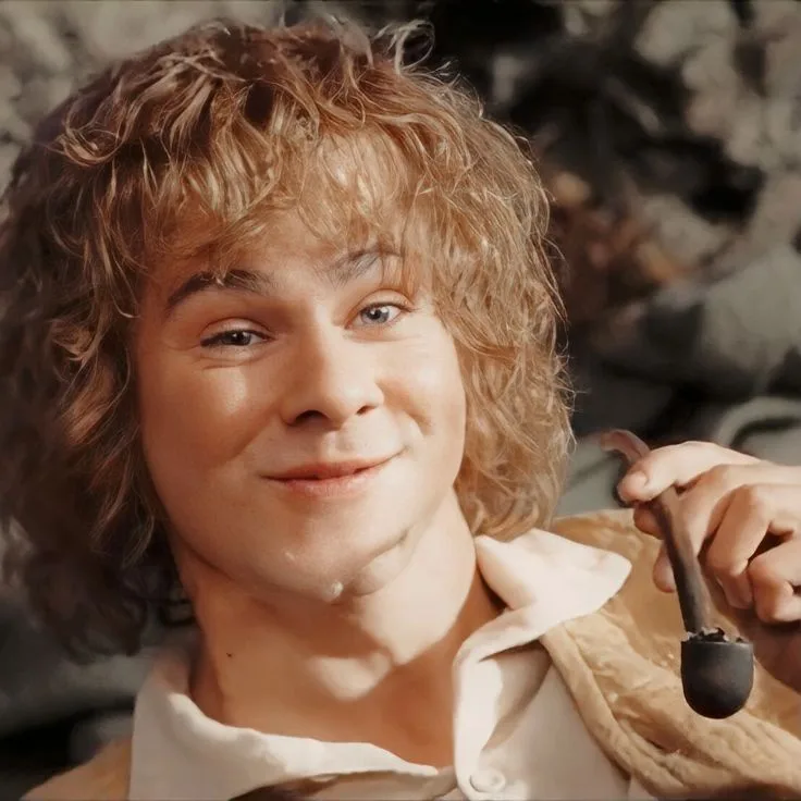 A close-up of a young man with curly blonde hair and a gentle smile, wearing a light-colored shirt and holding a pipe. The…