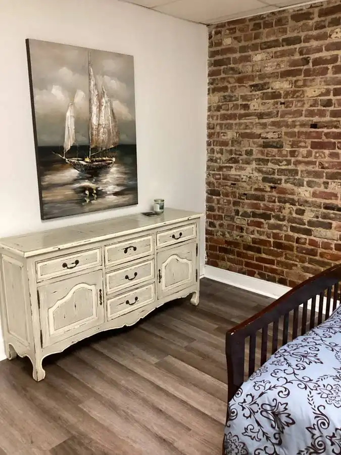 a bedroom corner showing a distressed white dresser against a white wall, with a large painting of a sailboat on the wall…