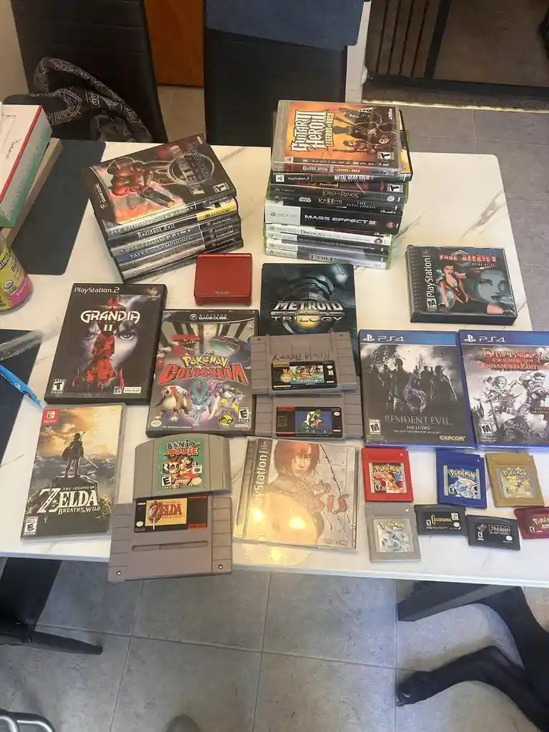 A white table displays a large collection of video games across multiple platforms, including physical cartridges for…
