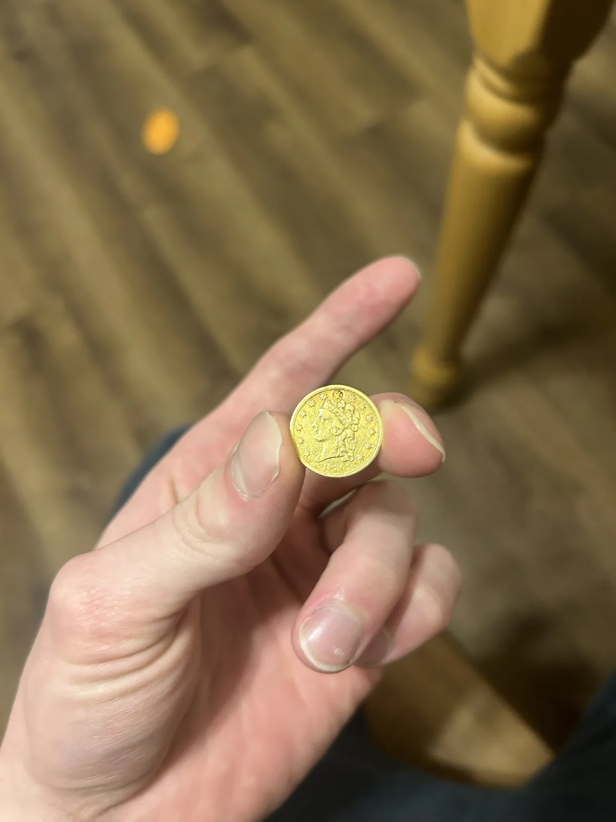 A person holds a small, gold-colored coin between their thumb and index finger, with a wooden floor and chair leg blurred in…