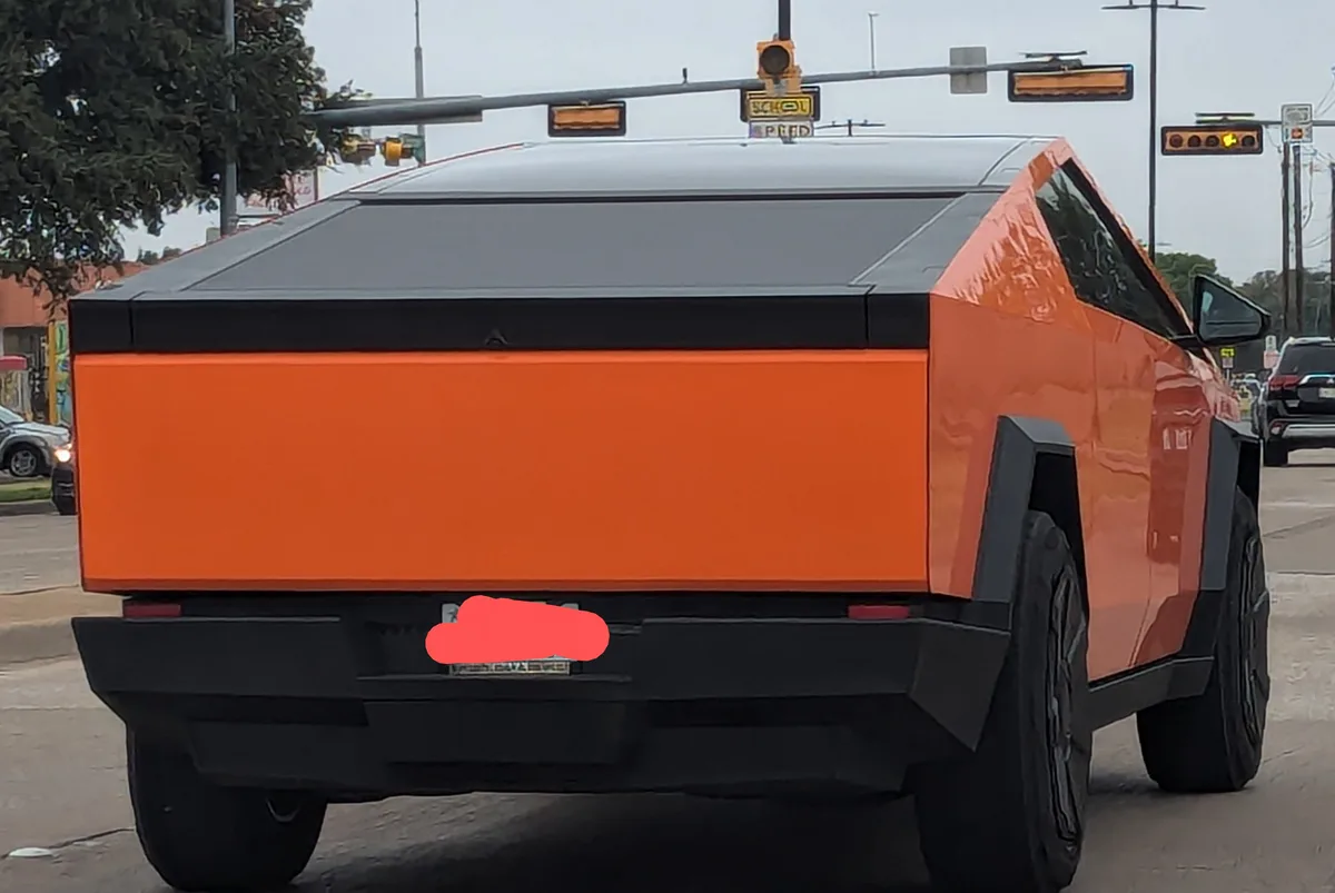 The image shows the rear three-quarter view of an orange and black Tesla Cybertruck at a traffic light. The vehicle's…