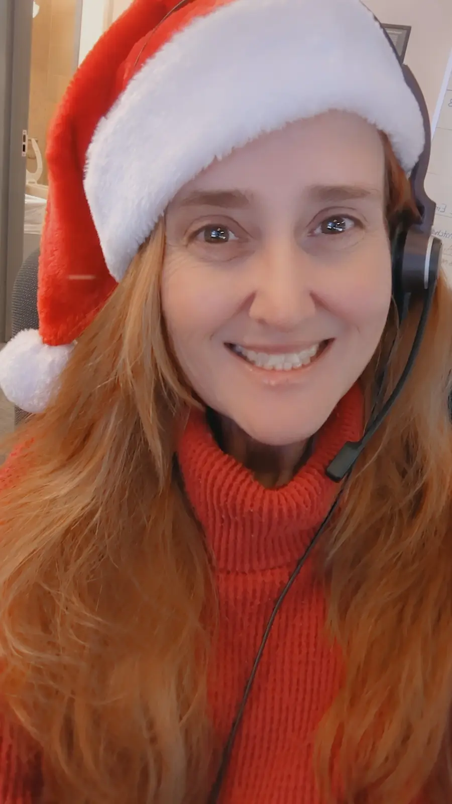 A woman with long red hair smiles at the camera while wearing a red and white Santa hat, a red turtleneck sweater, and a…