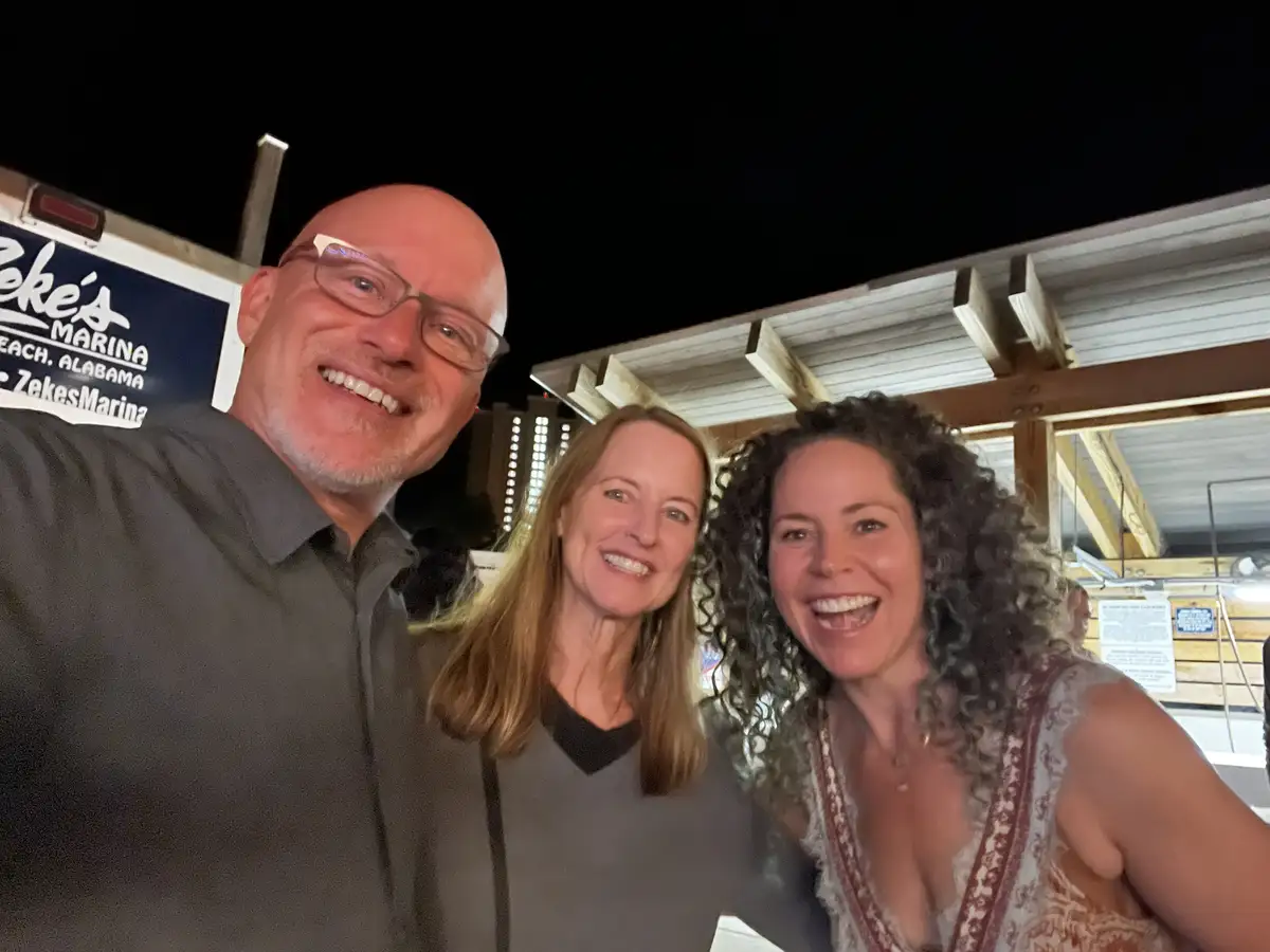 Three smiling people pose for a selfie at night in front of a sign that reads 'Zeke's Marina Beach, Alabama'. The man on the…