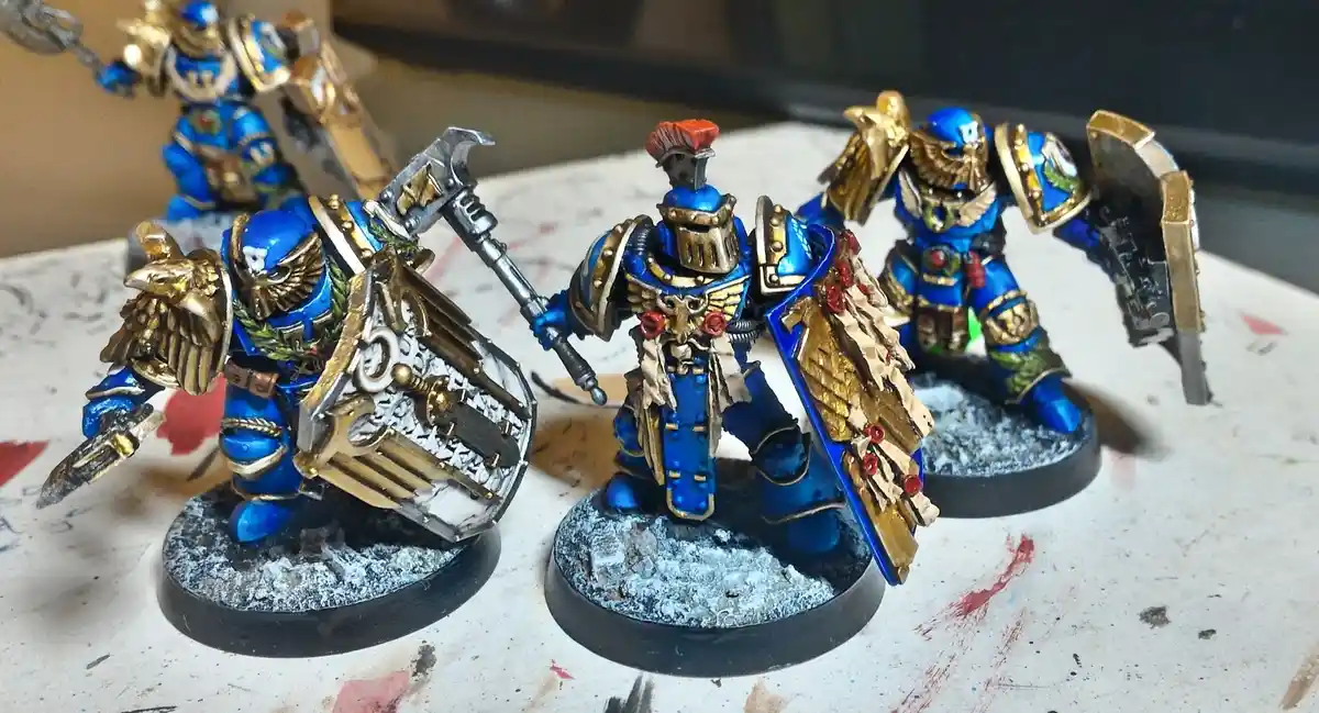 Three intricately painted blue and gold fantasy miniatures, resembling Space Marines, stand on bases with weathered, snowy…