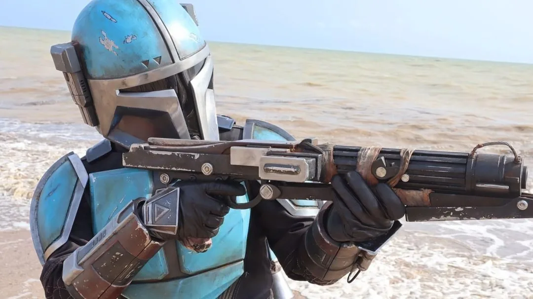 A person in a blue and silver Mandalorian-style helmet and armor stands on a beach, holding a blaster rifle with both hands…