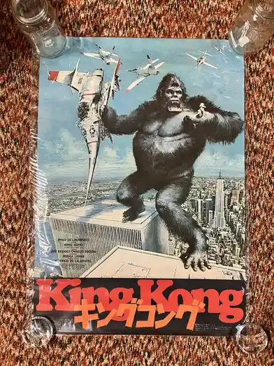A vintage Japanese movie poster for 'King Kong' featuring the giant ape holding a fighter jet aloft while standing atop a…