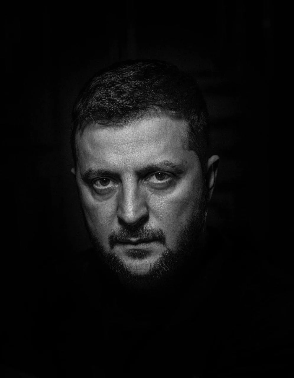 A black and white portrait of a man with a serious, intense gaze, set against a dark background. The image has text overlaid…