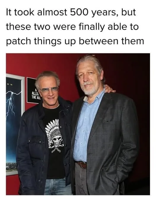 Two older men, one in a black t-shirt with a skull graphic and glasses, the other in a blue shirt and blazer with his arm…
