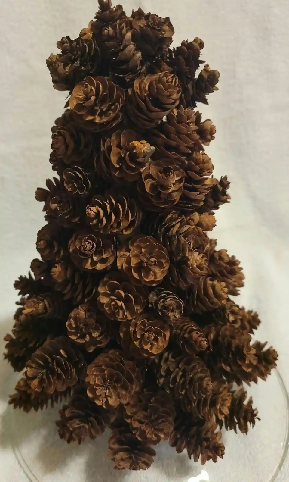 A decorative Christmas tree made entirely of brown pinecones arranged in a triangular shape, sitting on a clear glass plate…