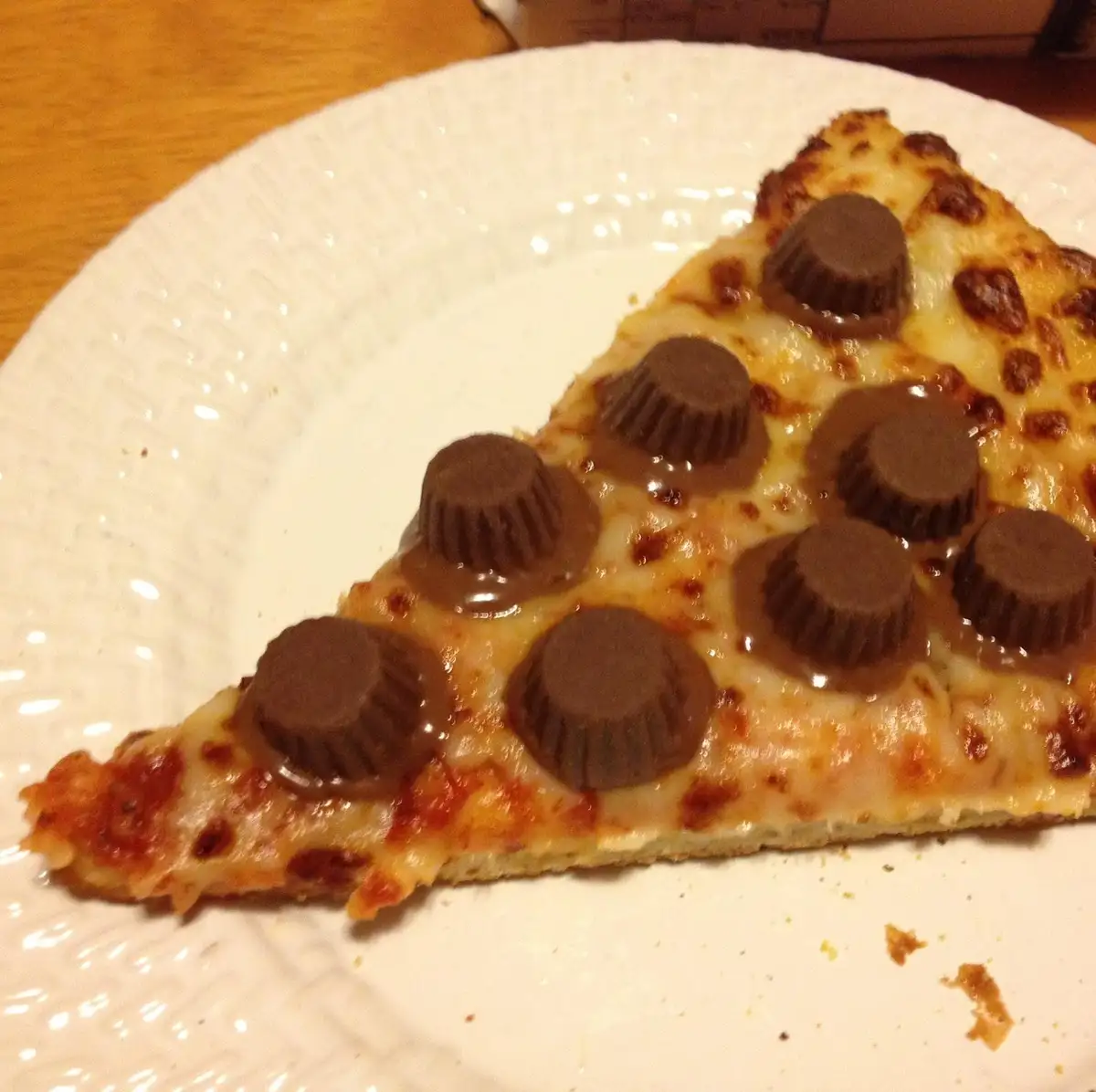 A slice of pizza with melted cheese and multiple peanut butter cups melted on top, served on a white paper plate. The text…