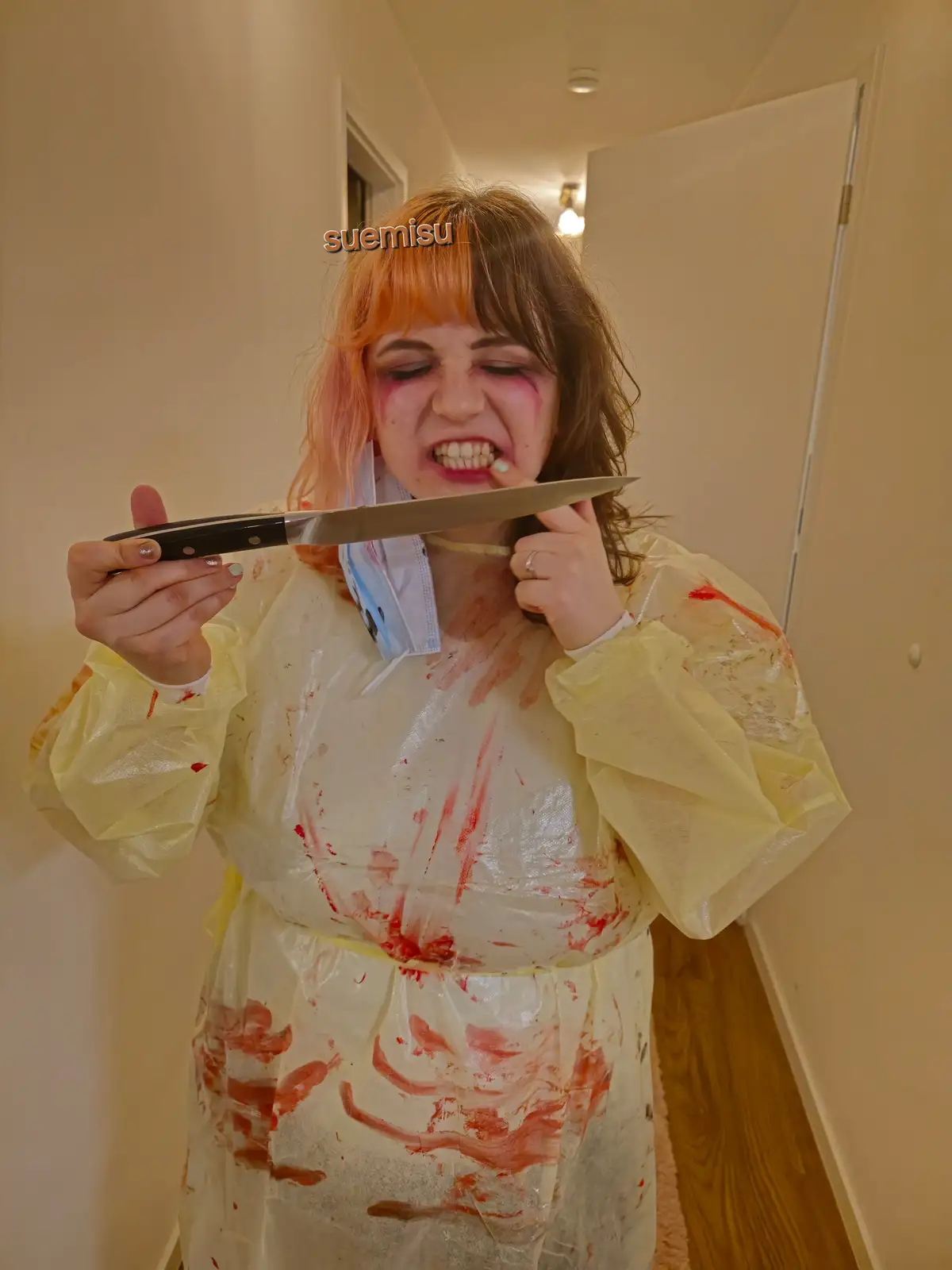 A person with orange hair and a yellow plastic raincoat covered in red paint or fake blood stands in a hallway, holding a…