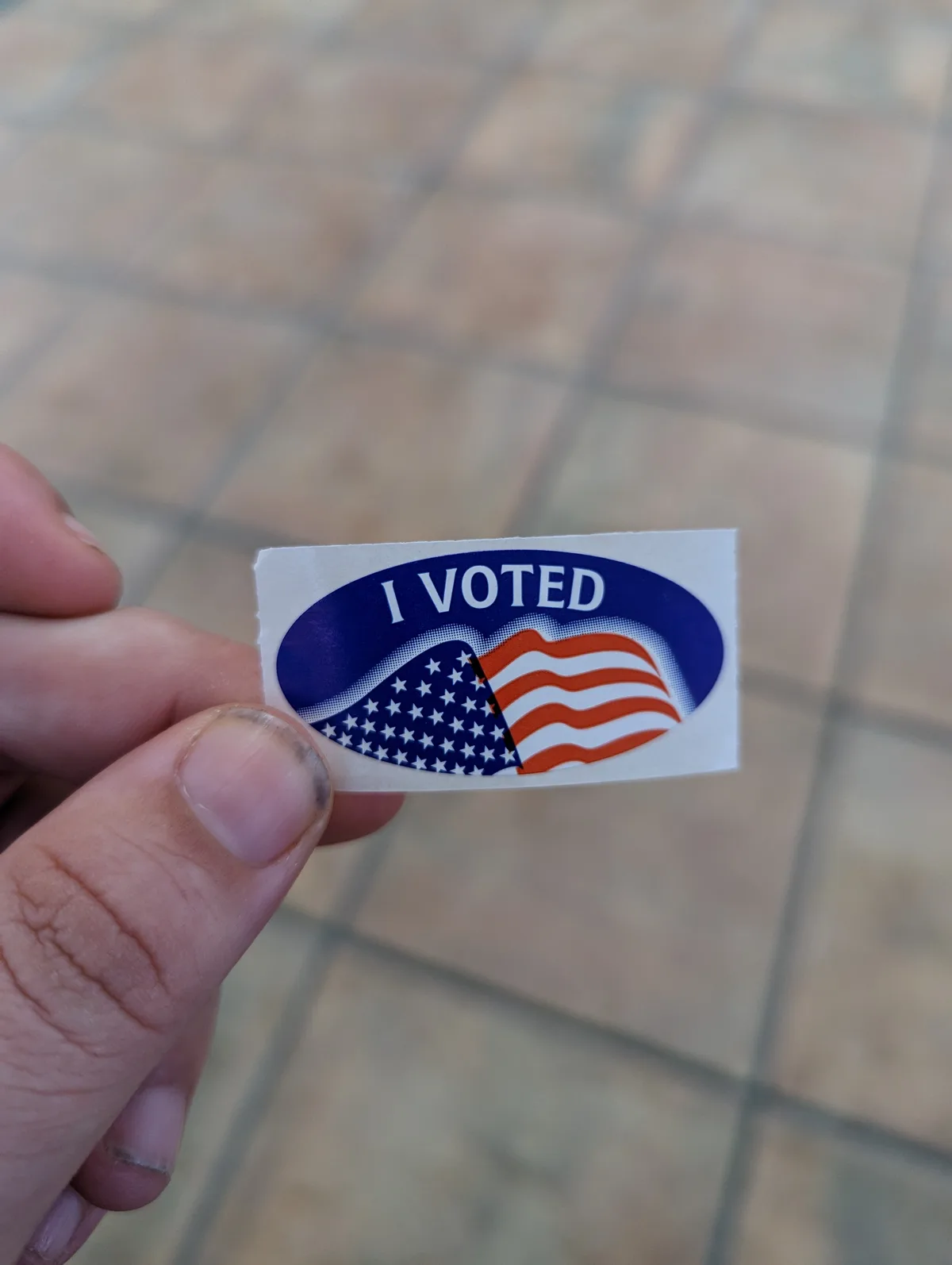 A hand holds a small, rectangular 'I VOTED' sticker featuring a stylized American flag design with stars and stripes, set…