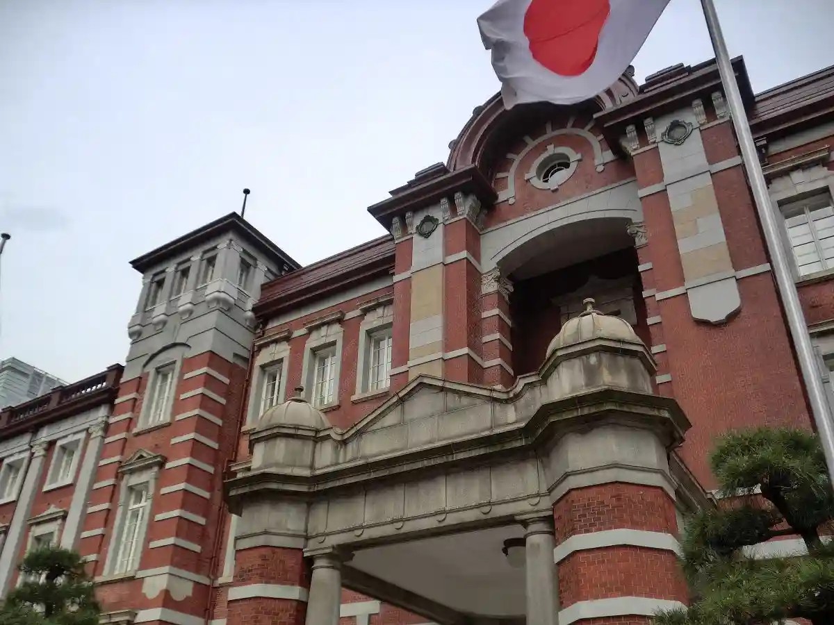 A low-angle photo of a red-brick Japanese government building with a Japanese flag waving above it, overlaid with the text…