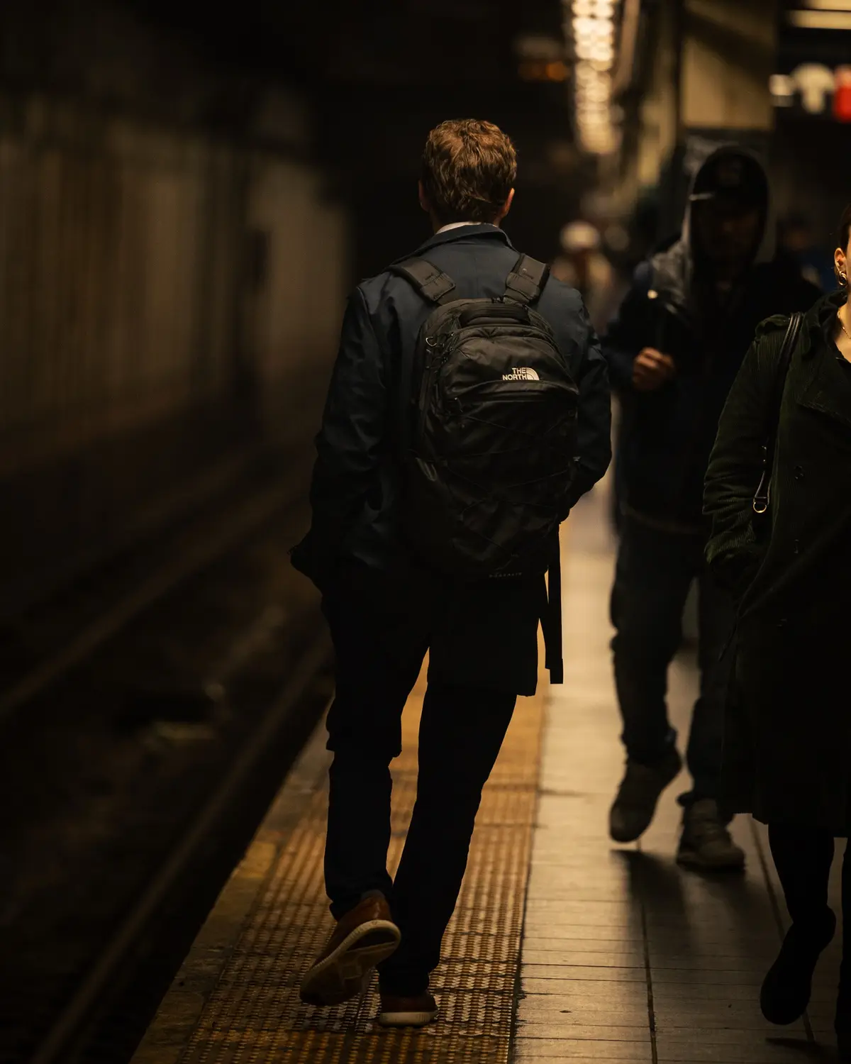 A man in a dark jacket and brown shoes walks away from the camera down a dimly lit subway platform, carrying a black…