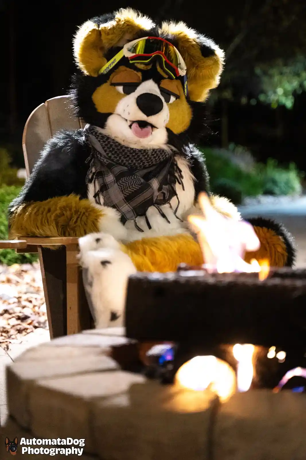 A person in a black, tan, and white furry costume resembling a dog sits in a wooden chair at night, wearing ski goggles…