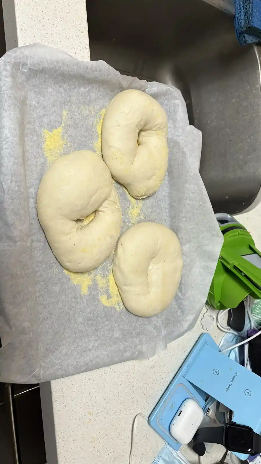 Three raw bagel doughs shaped like bagels sit on parchment paper next to a sink, with yellow flour dusting the paper. The…