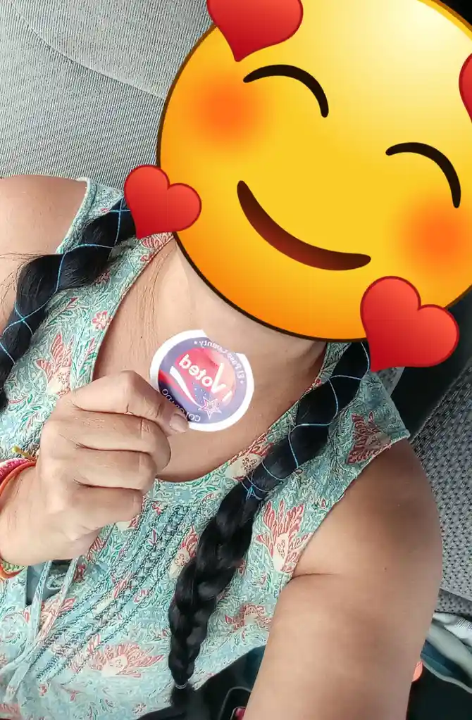 A person with braided hair and a patterned top holds up an 'I Voted' sticker on their chest, while their face is obscured by…