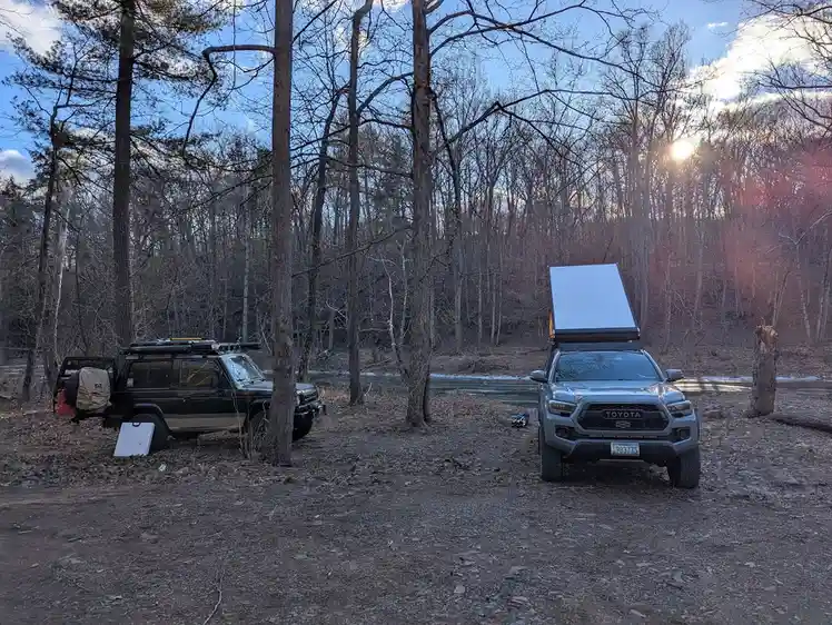 Two off-road vehicles are parked in a wooded area near a river at sunset. A silver Toyota Tacoma with a rooftop tent is…