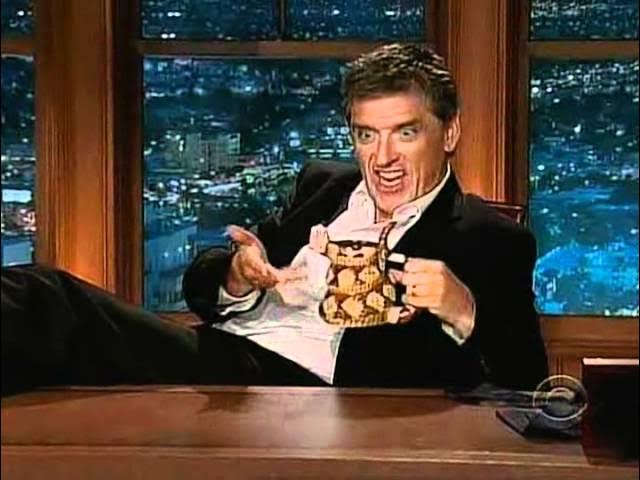 a man in a suit sits at a desk holding a mug with a patterned design, his face contorted in an exaggerated expression of…