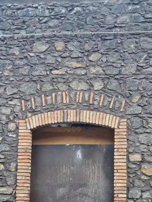 A close-up of a rustic stone wall with a wooden door framed by red bricks. The brick arch above the door is unevenly laid…
