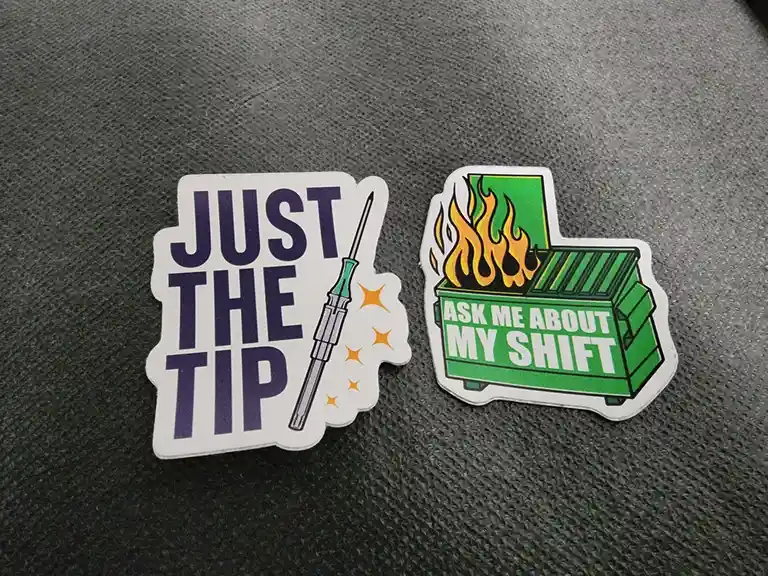 Two stickers lie on a gray textured surface. The left sticker has purple text reading 'JUST THE TIP' next to a screwdriver…