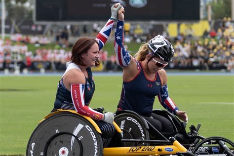 Two female wheelchair racers in athletic gear celebrate together on a track, raising their clasped hands high in victory…