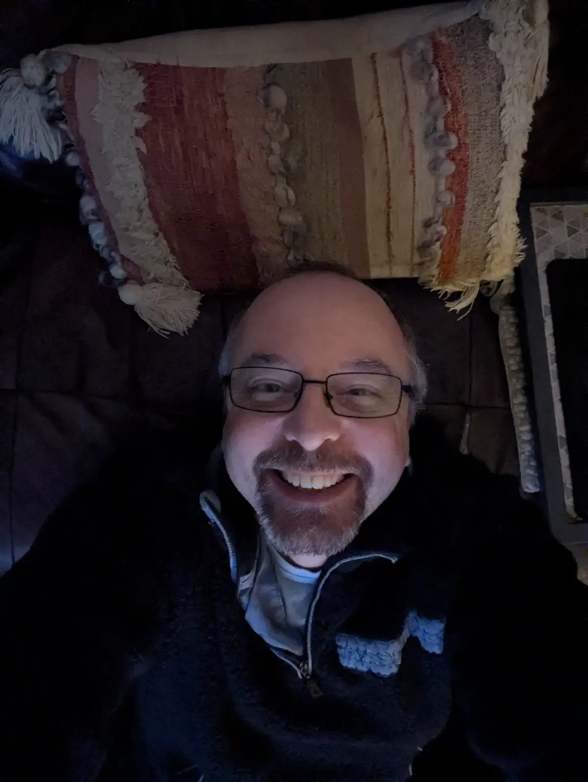 A man with glasses and a goatee smiles broadly while lying on a couch, with a striped, tasseled pillow behind his head. The…