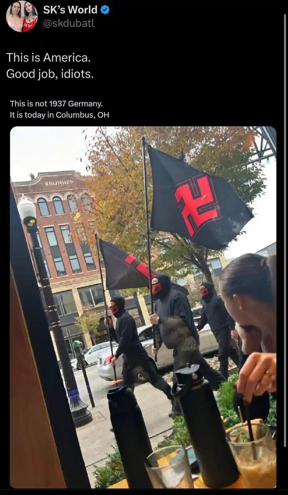 A photo taken from inside a cafe shows several masked individuals marching down a city street while carrying black flags…