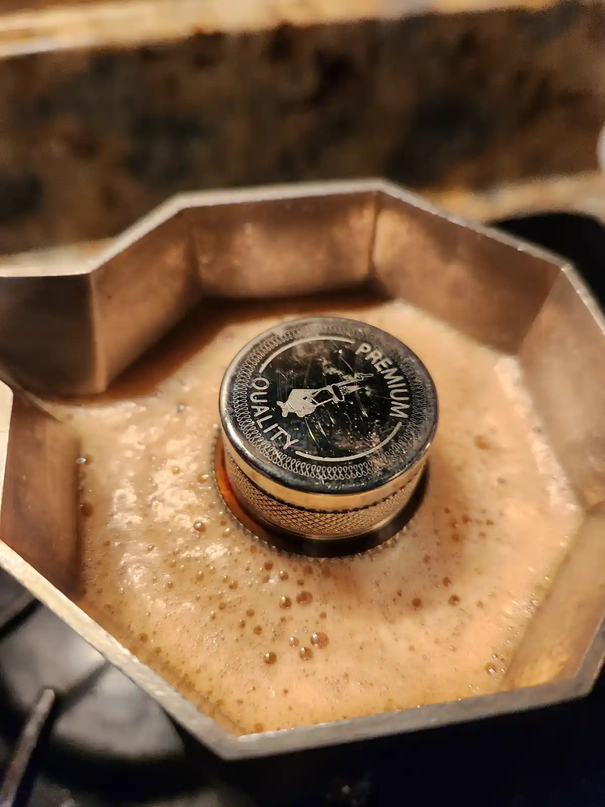 A close-up of a used moka pot with coffee foam surrounding its central knob, which has the words 'PREMIUM QUALITY' engraved…