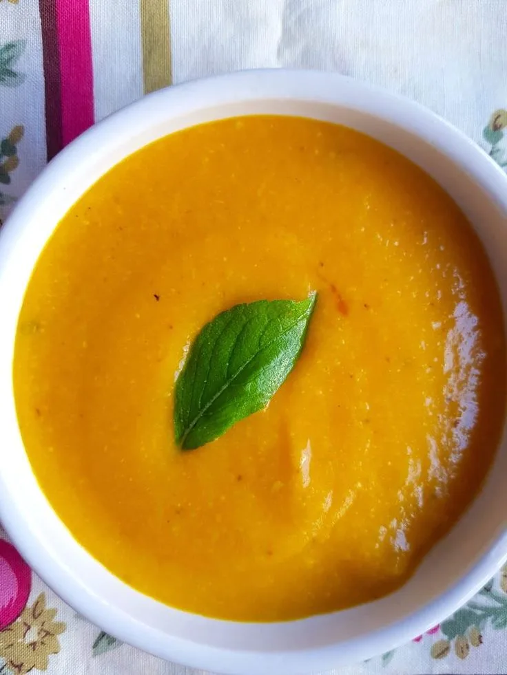 A top-down photo shows a white bowl filled with smooth, orange-colored soup, garnished with a single green leaf resting in…