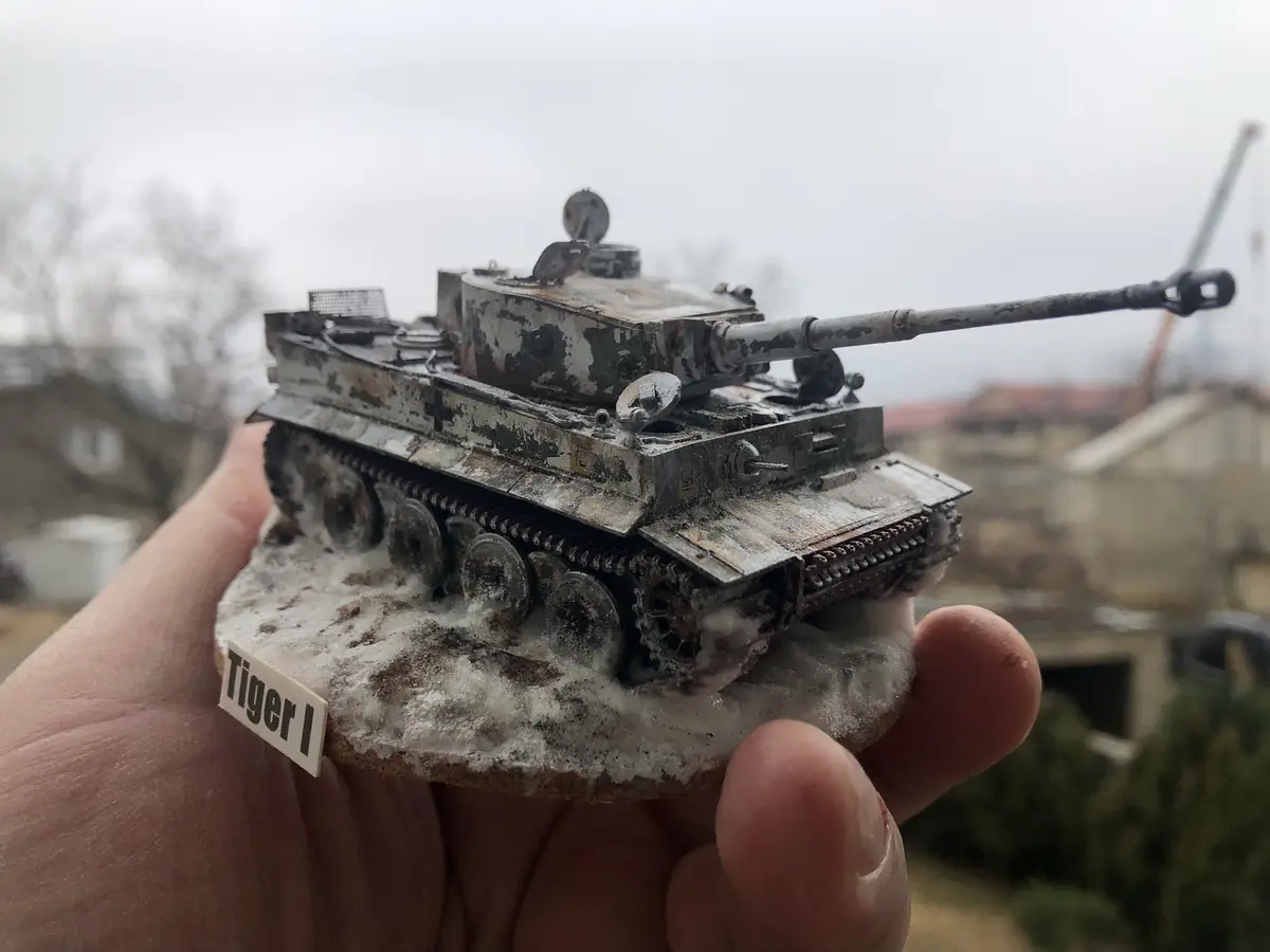 A hand holds a highly detailed, weathered miniature model of a Tiger I tank, which is mounted on a base resembling a snowy…