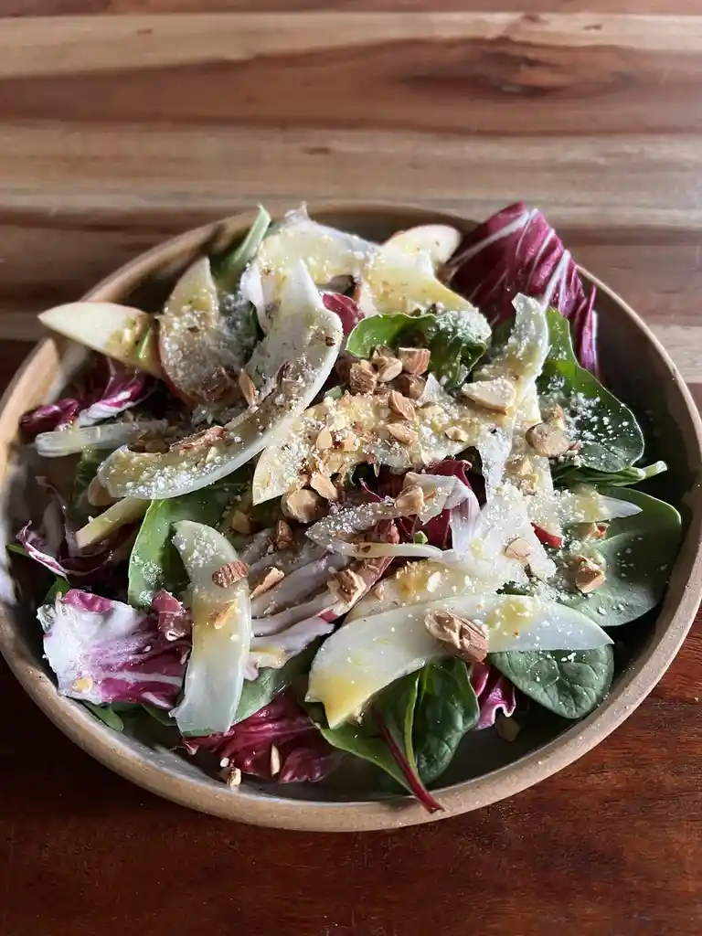 A close-up photo of a colorful salad in a wooden bowl, featuring mixed greens, thinly sliced apples, shaved cheese, chopped…