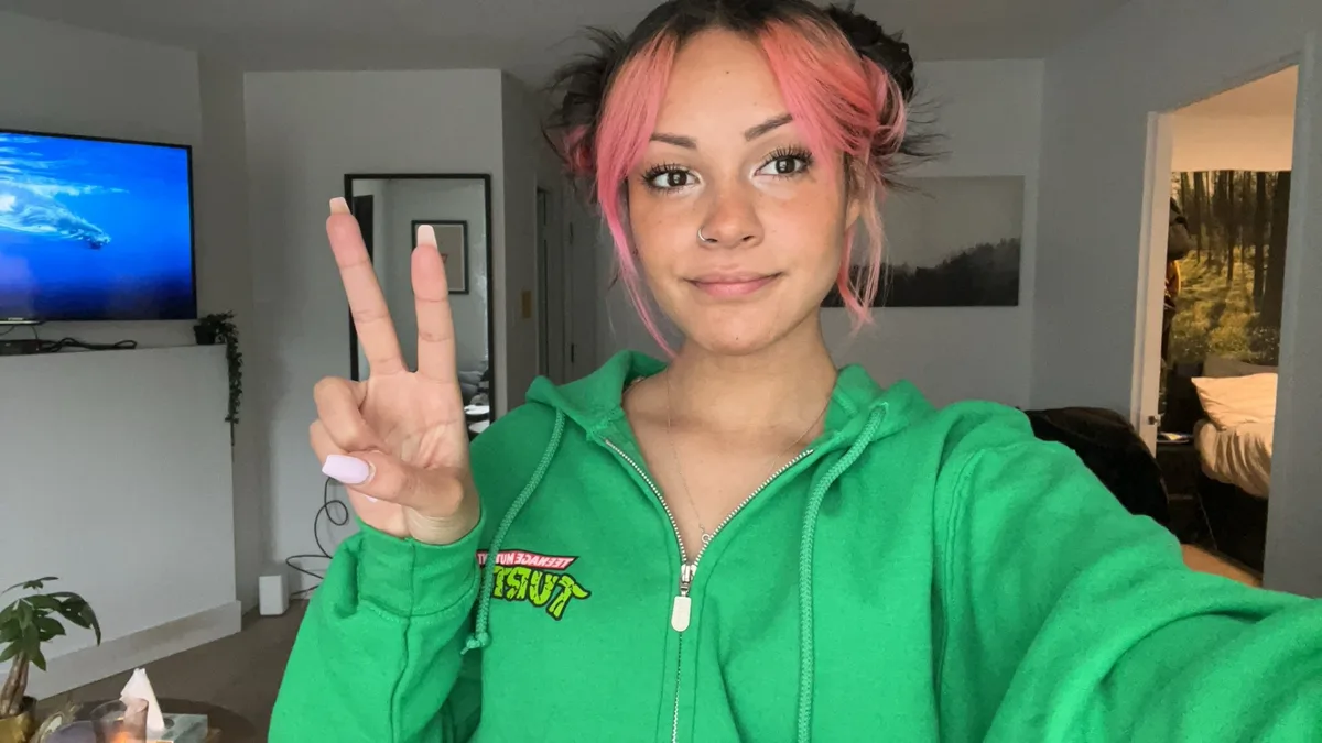 A young woman with pink and black hair styled in buns, wearing a green Teenage Mutant Ninja Turtles hoodie, holds up a peace…