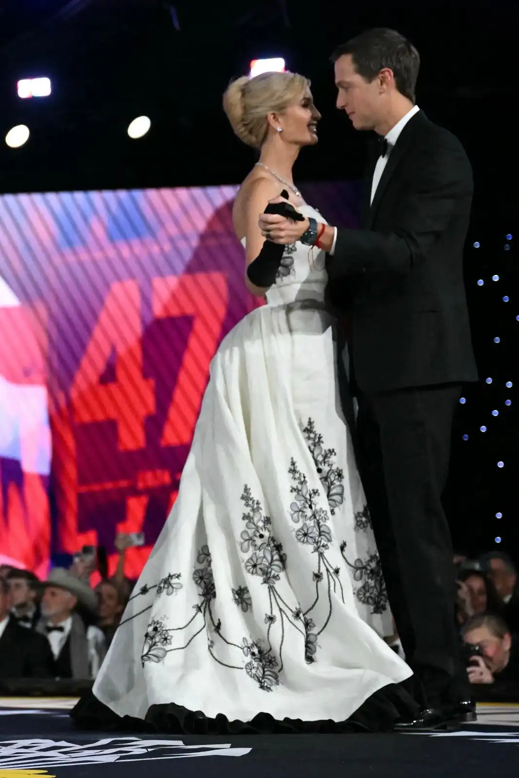 A man and woman are standing close together on a stage, dressed formally for an event. The woman wears a white gown with…