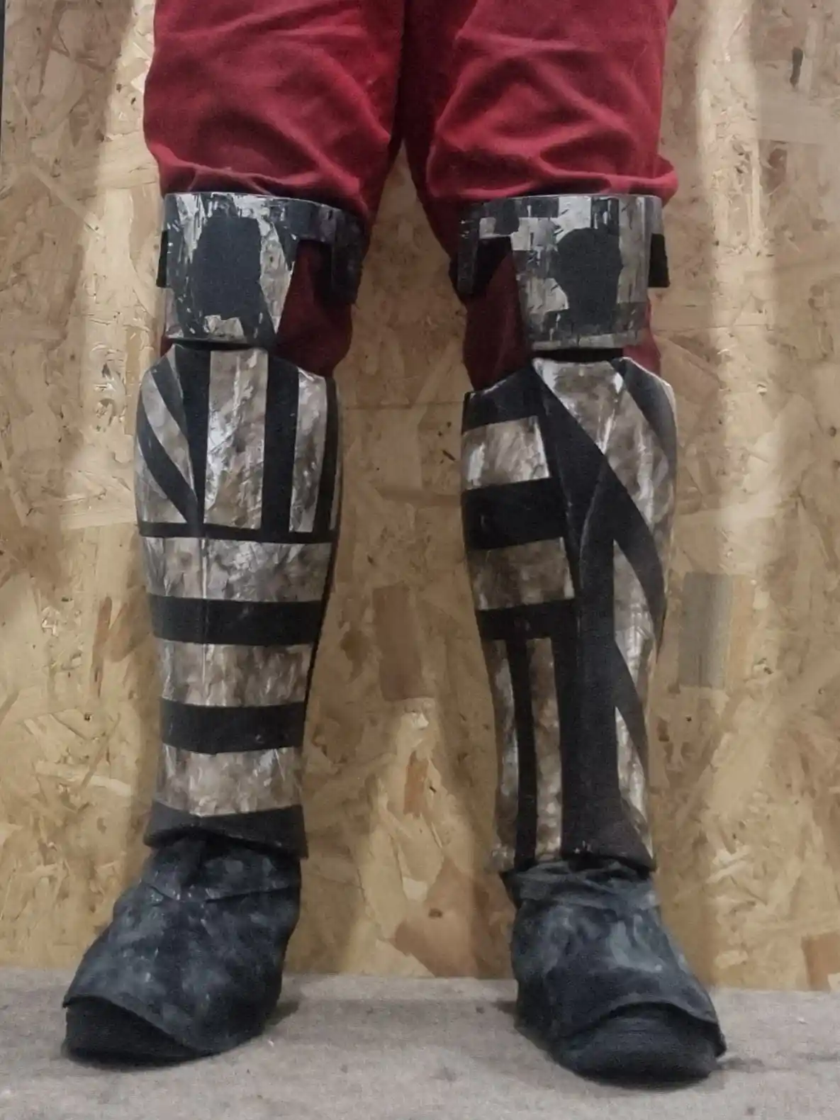 A person wearing red pants and homemade, heavily patched, and distressed-looking cosplay leg armor with black and silver…