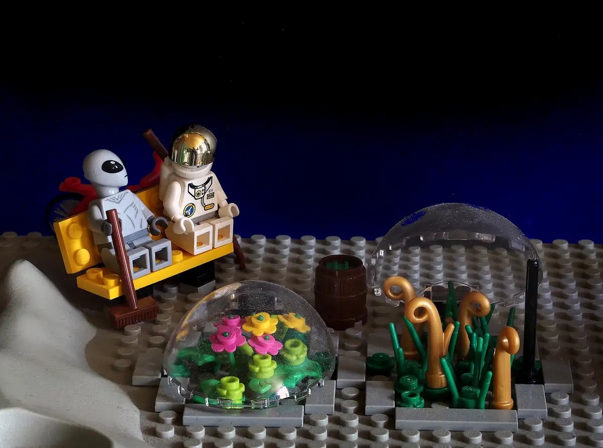 A LEGO diorama shows two minifigures, one alien and one astronaut, sitting on a makeshift bench made of LEGO bricks. In…