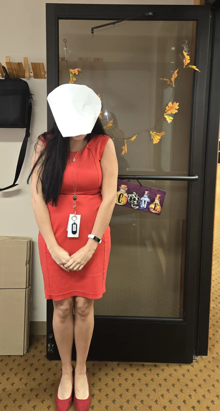 A woman in a red dress and red heels stands with her hands clasped in front of her, wearing a white paper bag over her head…